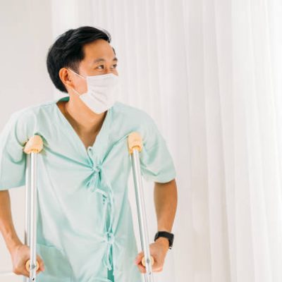 istockphoto-1320924590-612×612 Young male Asian patient in uniform walking with crutch walk in hospital while wearing a face mask